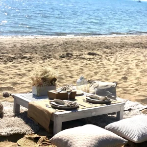 Serene beach picnic setup melbourne Serene beach picnic setup in Melbourne with coastal styling, low white table, neutral cushions, and dried floral centerpiece